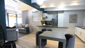 Un salon moderne avec un canapé gris, une table à manger avec des chaises, un coin cuisine avec des meubles blancs et une grande fenêtre avec des rideaux.