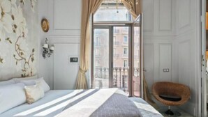 Une chambre d’hôtel avec un grand lit, une chaise en osier et une vue sur les immeubles par la fenêtre.