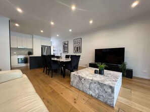A modern living room with a marble coffee table, a sofa, a dining area with chairs, a kitchen with cabinets, and a flat-screen TV.