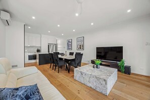 Ein modernes Wohnzimmer mit einer Couch, einem Marmortisch, einem Essbereich mit Stühlen, einer Küche mit weißen Schränken und einem Flachbildfernseher.