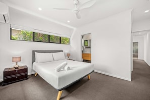 Ein modernes Schlafzimmer mit einem großen Bett, einem Nachttisch, einer Kommode, einem Deckenventilator und einem Fenster mit Blick auf Grünflächen.