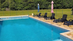Ein rechteckiger Swimmingpool mit gefliestem Rand, umgeben von einer Steindeck. Auf der Deck liegen schwarze Liegestühle und ein pinkfarbener Sonnenschirm.