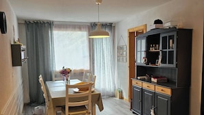 Ein Essbereich mit Holztisch und Stühlen, einer Hängeleuchte, einem Schrank mit geöffneten Türen und einem Fenster mit Vorhängen.