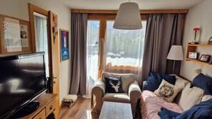 Ein gemütliches Wohnzimmer mit Fernseher, Holzmöbeln, einer Couch mit dekorativen Kissen, einem Couchtisch und Blick auf eine verschneite Landschaft durch große Fenster.