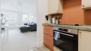 Une cuisine moderne dotée d’un four encastré, un coin repas avec une table et des chaises blanches, et un salon avec un canapé et une plante.