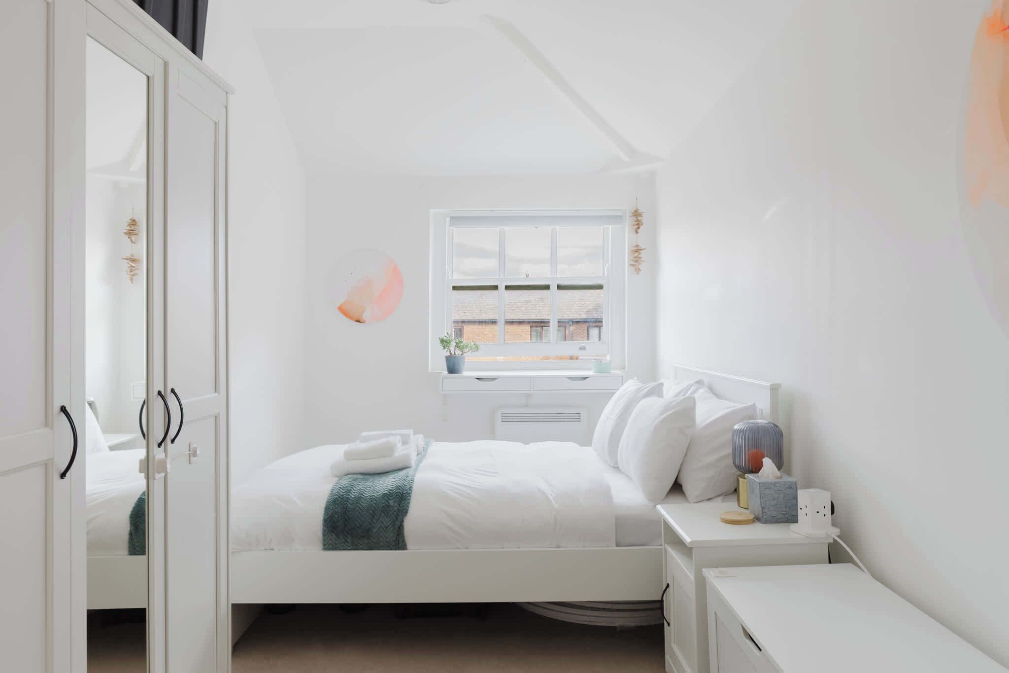 A bedroom with a bed, bedside table, nightstand, wardrobe, window, and a view of the outside.