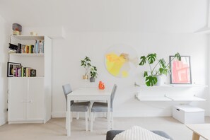 Una habitación moderna con un escritorio blanco, sillas, una estantería, una planta en maceta y un cuadro en la pared.