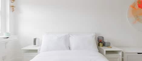A minimalist hotel room with a large bed, white bedding, a bedside table with a lamp, and a wall-mounted air purifier.