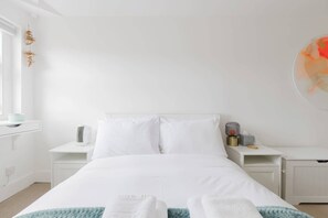 A white bed with pillows, a bedside table with a lamp, and a wall-mounted artwork.