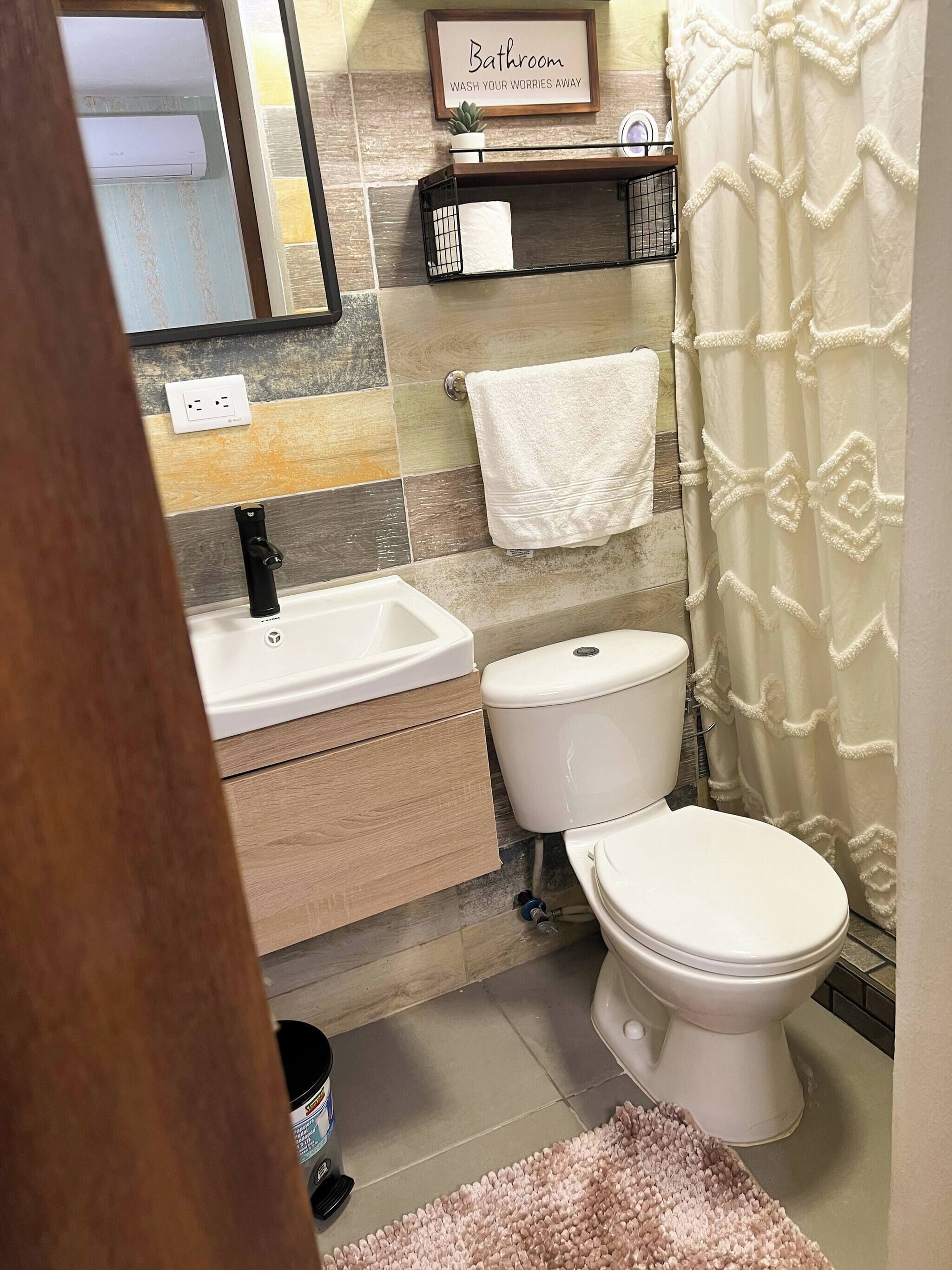 Ein Badezimmer mit weißem Waschbecken, Toilette und Duschvorhang. Ein Handtuch hängt an der Wand und ein kleines Regal mit einer Pflanze ist vorhanden.