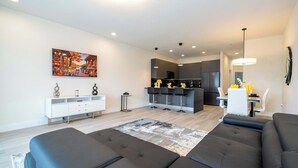 A modern living room with a sofa, a dining area with a table and chairs, and a kitchen in the background. - Modern 3Bd Screened Pool at Serenity Resort 17322 (Clermont)