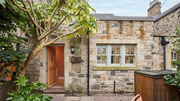 A stone cottage with a wooden door, a window with white frames, and a small garden area with a tree and a bench. - Host Stay Sonsy Cottage (Alnwick)