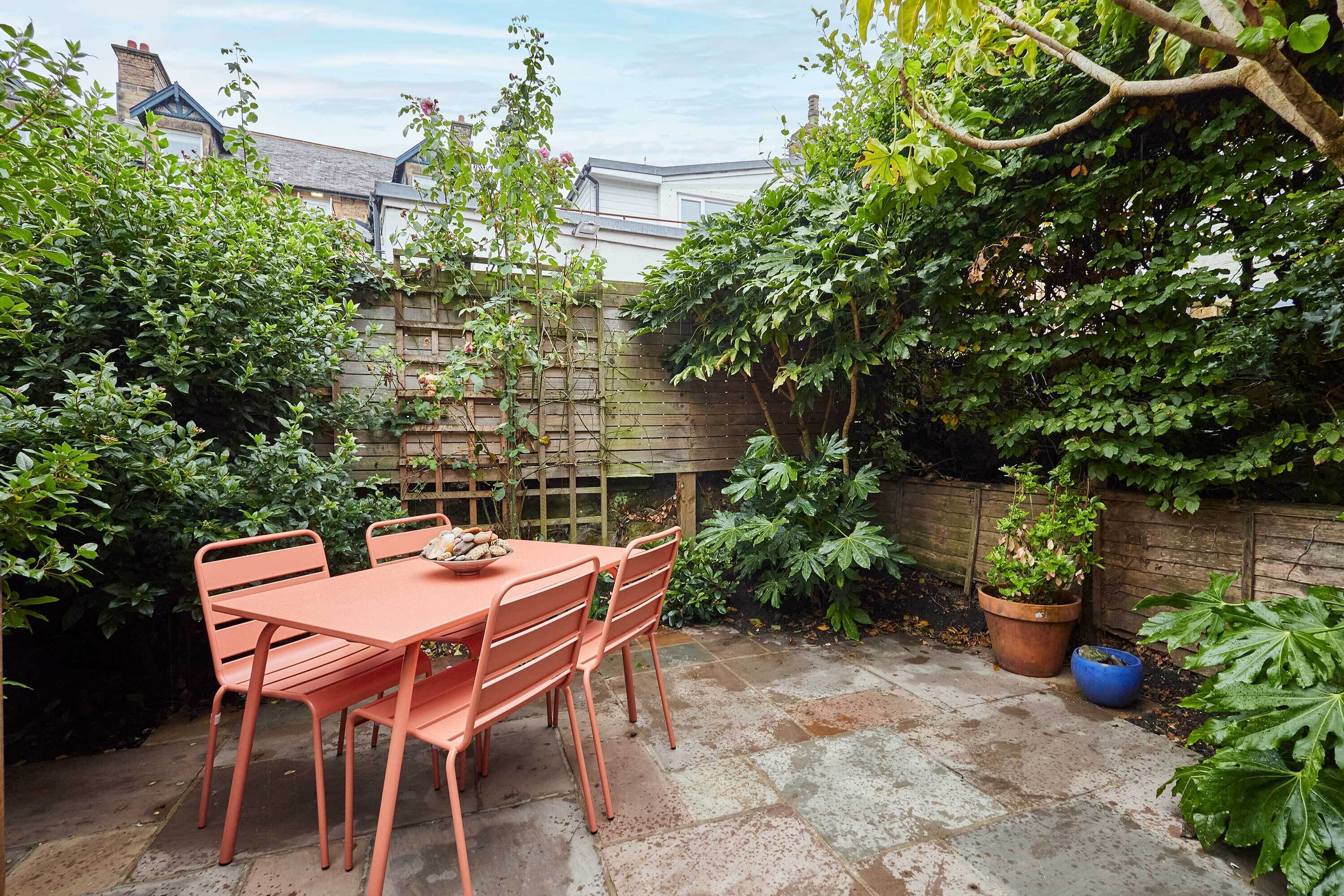 A patio with a table and chairs, surrounded by lush greenery and a wooden fence.