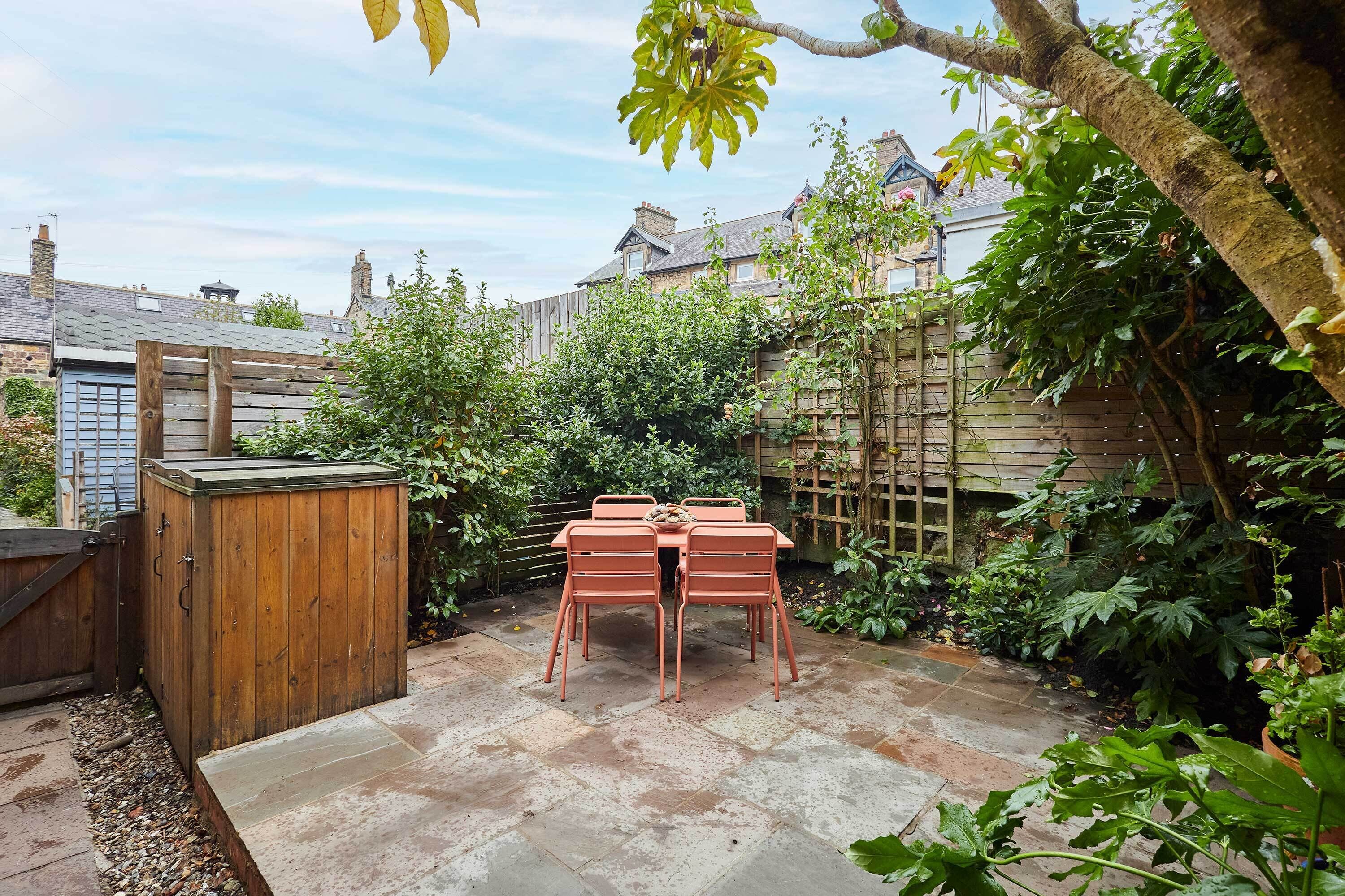A patio with a wooden table and chairs, surrounded by a garden with various plants and a wooden fence.