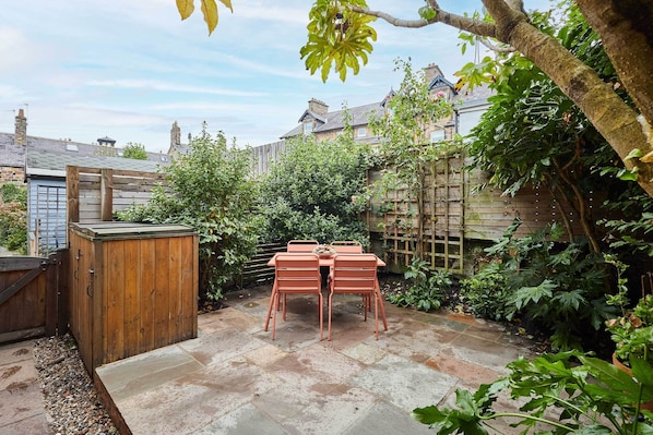 A patio with a wooden table and chairs, surrounded by a garden and a wooden fence.