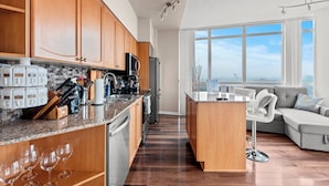 A modern kitchen with wooden cabinets, a granite countertop, and stainless steel appliances. There is a dining area with a gray sofa and a large window offering a view of the city. - Atlantis at Waterpark City Condos in DT (Toronto)