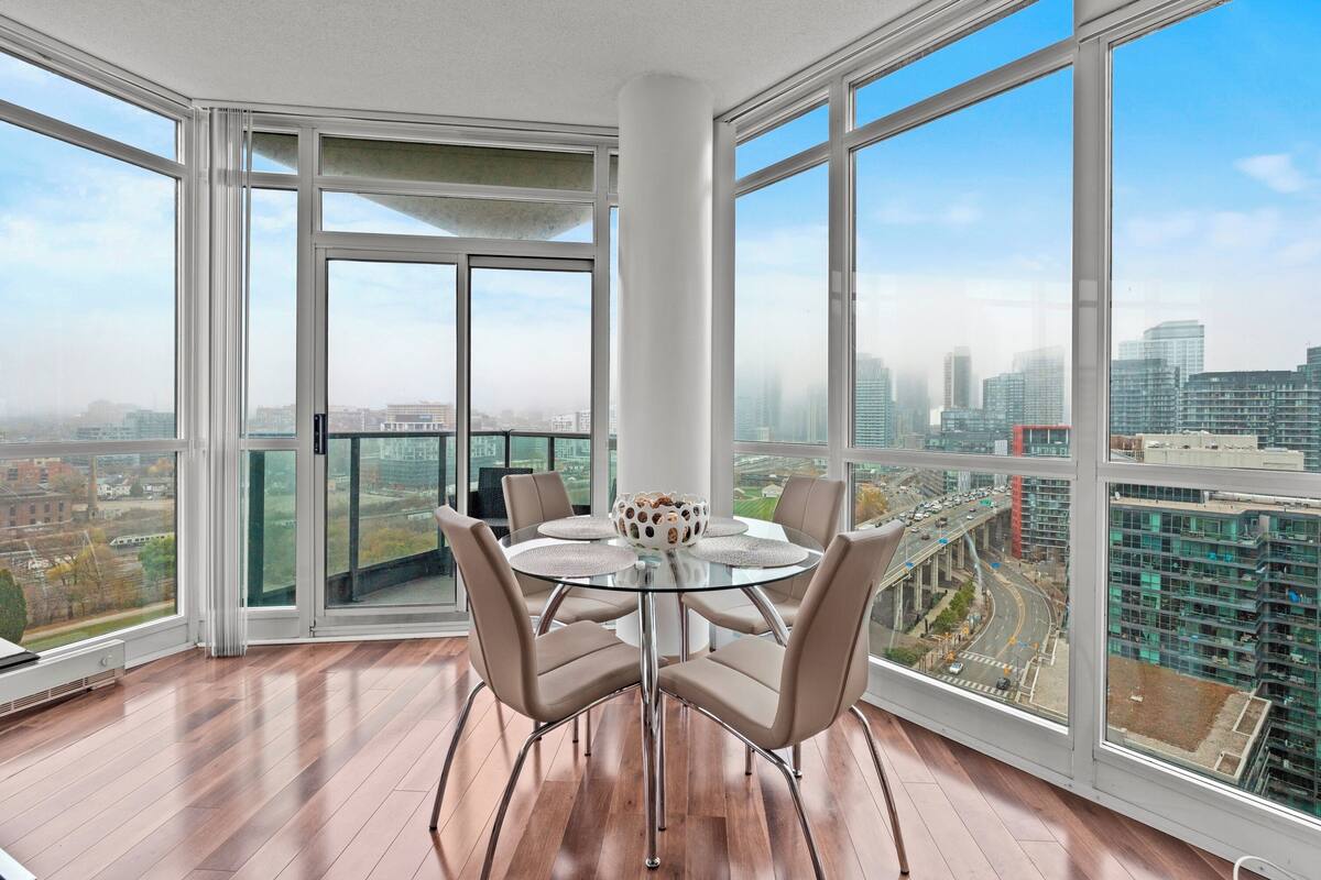 A modern dining area with a glass table and chairs, large windows offering city views, and a white pillar.