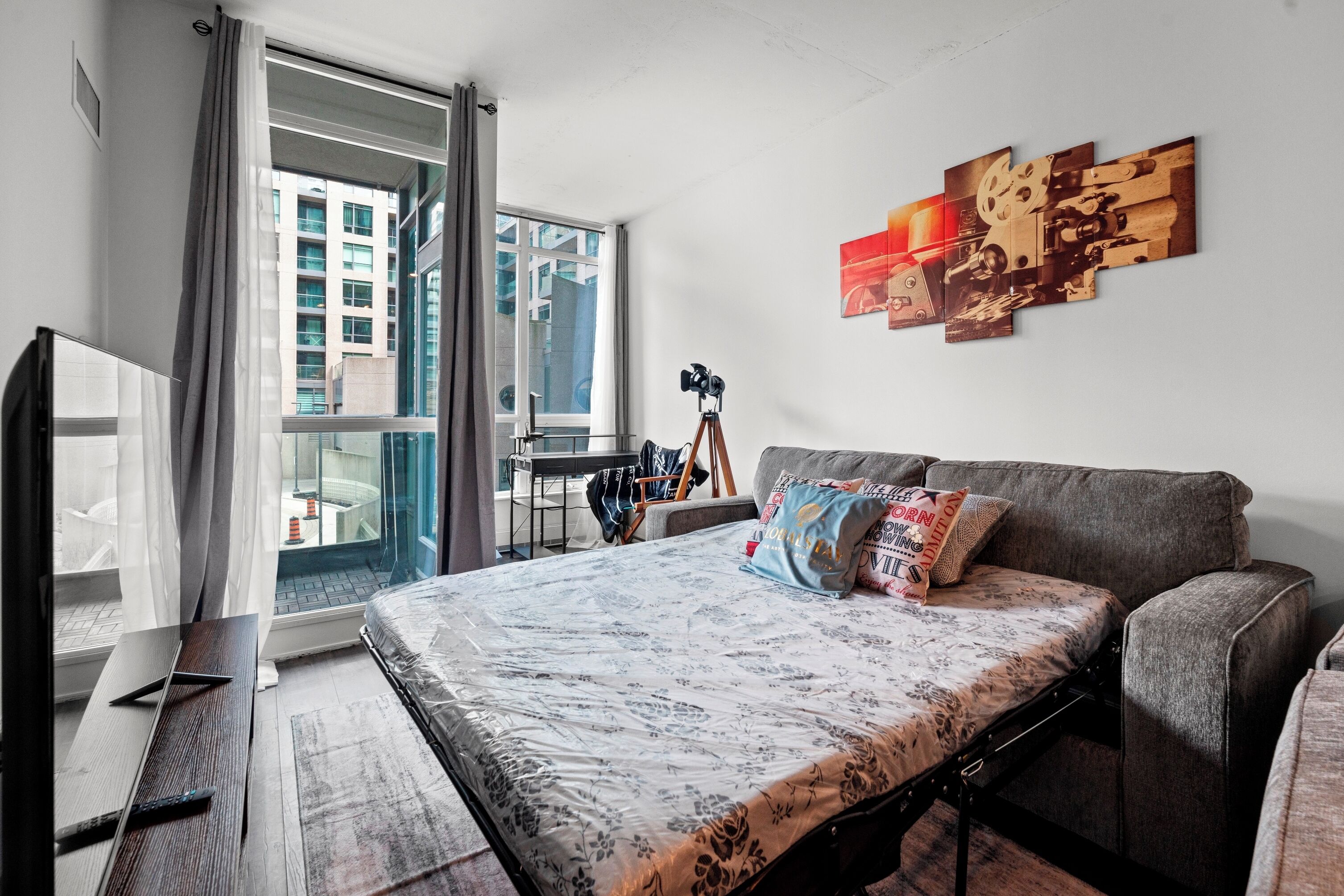 A modern bedroom with a large bed, a grey sofa, a desk with a camera, and a window with curtains.