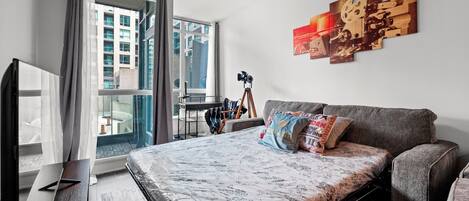 A modern bedroom with a large bed, a grey sofa, a desk with a camera, and a window with curtains.