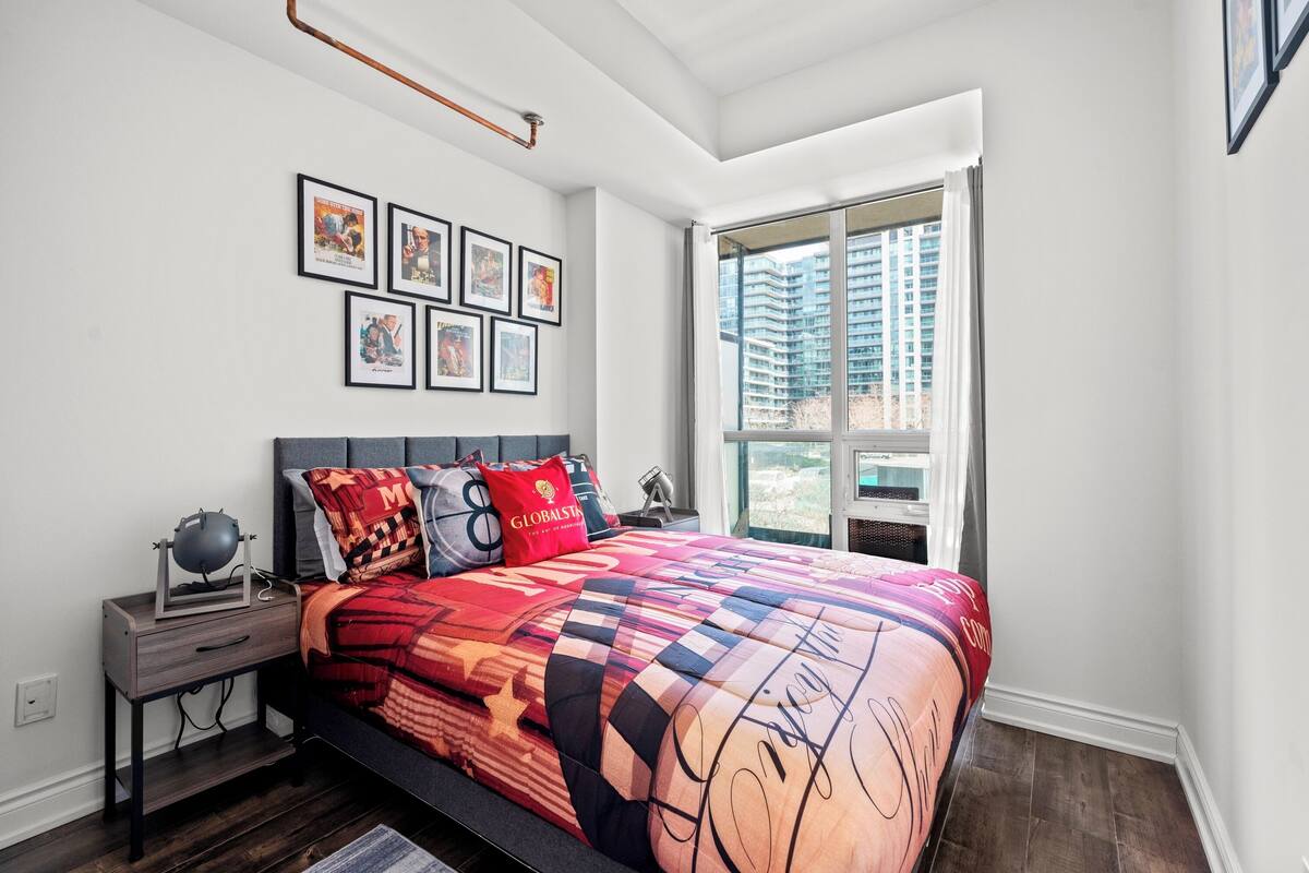 A bedroom with a large bed, a nightstand, a lamp, and a window with a view of high-rise buildings.