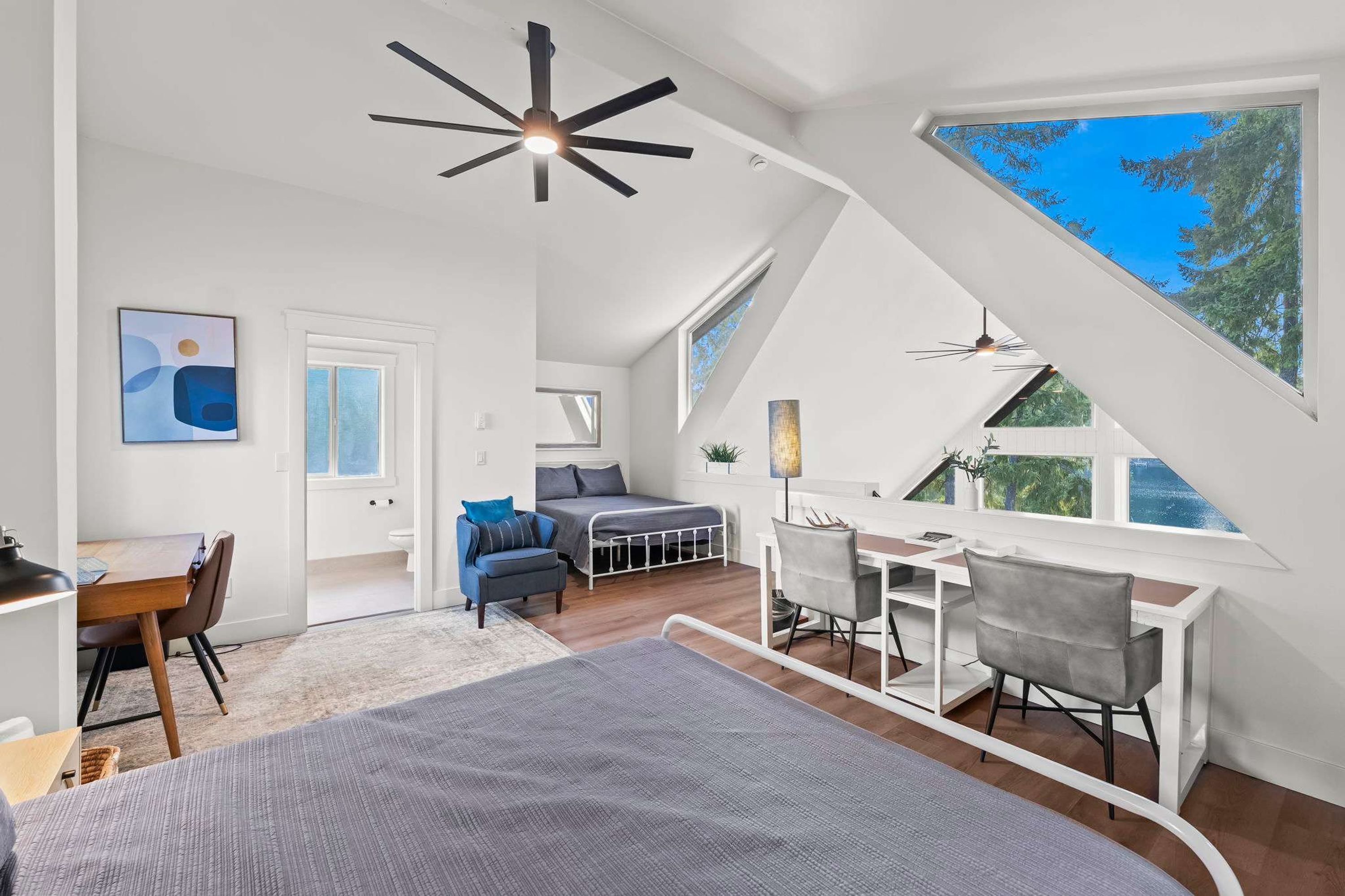 A modern loft bedroom with a sloped ceiling, a desk, and a large window offering a view of the outdoors.