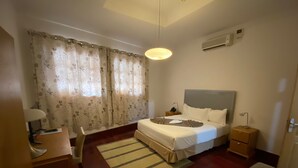 A hotel room with a bed, bedside tables, a desk, a chair, a lamp, a rug, and a window with curtains. - Mar... Guesthouse (Lobito)