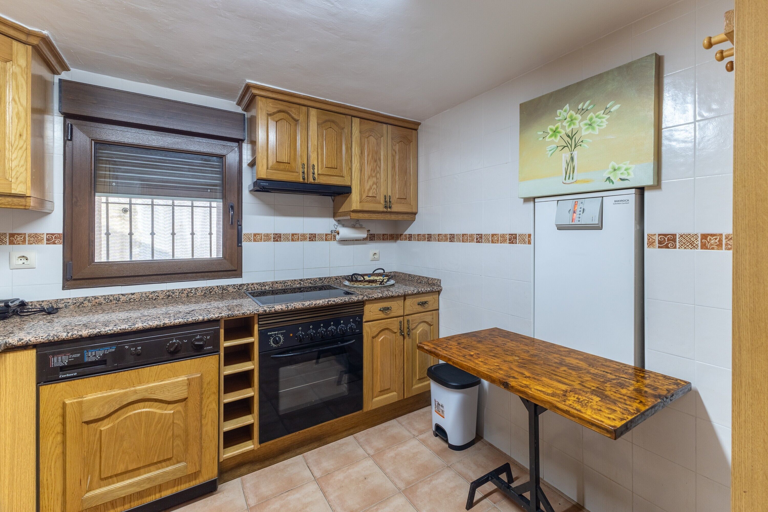 A kitchen with wooden cabinets, a built-in oven, a dishwasher, a window with blinds, a painting of flowers, and a refrigerator.