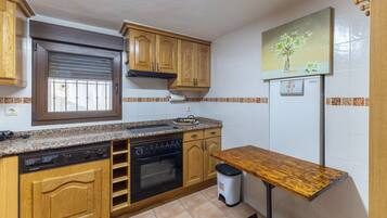 A kitchen with wooden cabinets, a built-in oven, a dishwasher, a window with blinds, a painting of flowers, and a refrigerator.