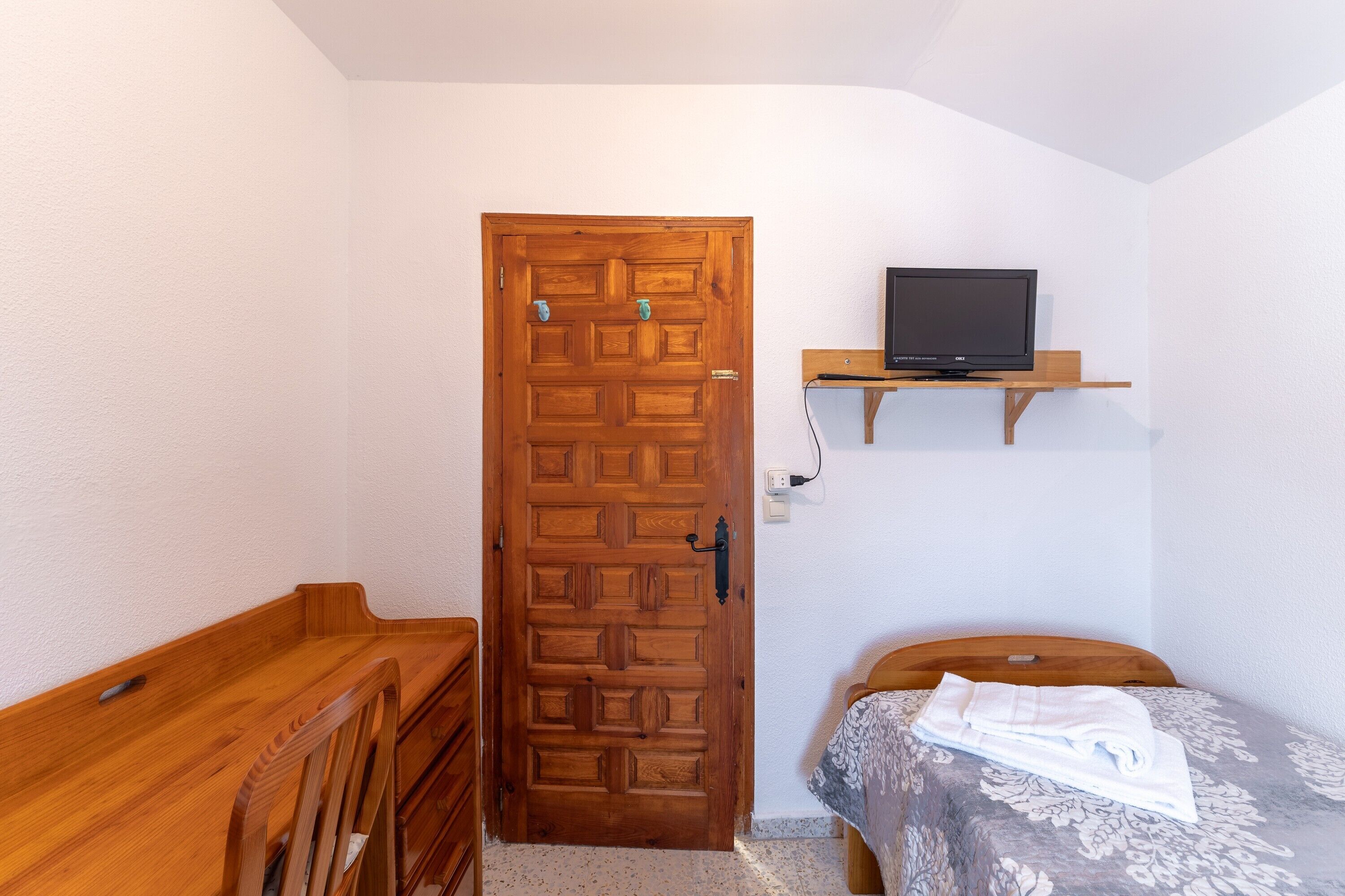 Une pièce avec une porte en bois, un lit recouvert d’une couvre-lit à motifs, un banc en bois et une étagère sur laquelle se trouve un téléviseur.