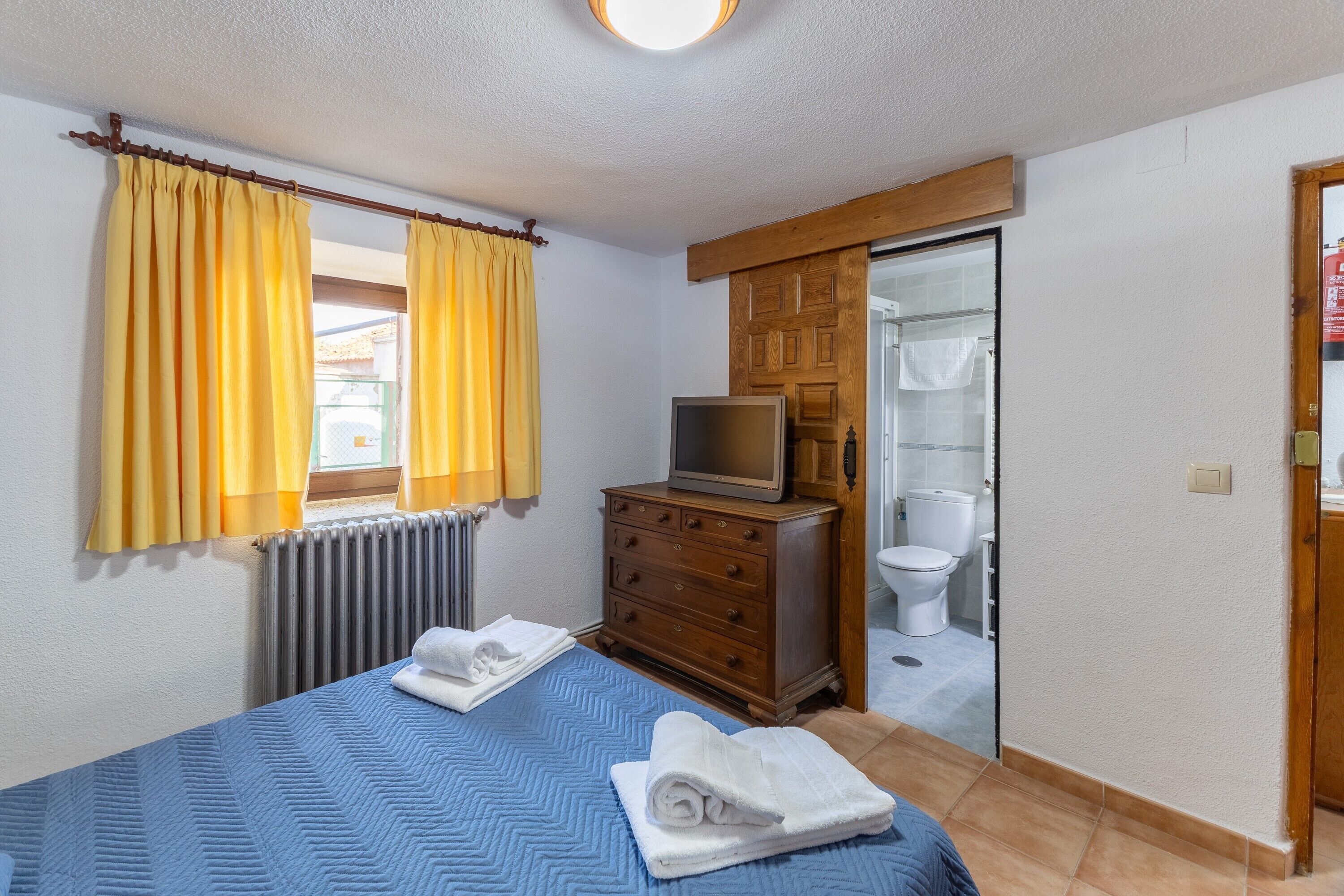 Un dormitorio con una cama, un aparador, un televisor y un baño visible a través de una puerta abierta.