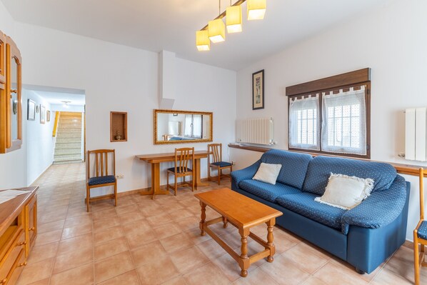 Un salon avec un canapé bleu, une table basse en bois et des fauteuils. On y trouve également une table à manger avec des chaises, un miroir et une fenêtre avec des rideaux.