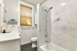Ein modernes Badezimmer mit Waschbecken, Toilette und Badewanne in Weiß, Marmorfliesen und einem Fenster mit Jalousien.
