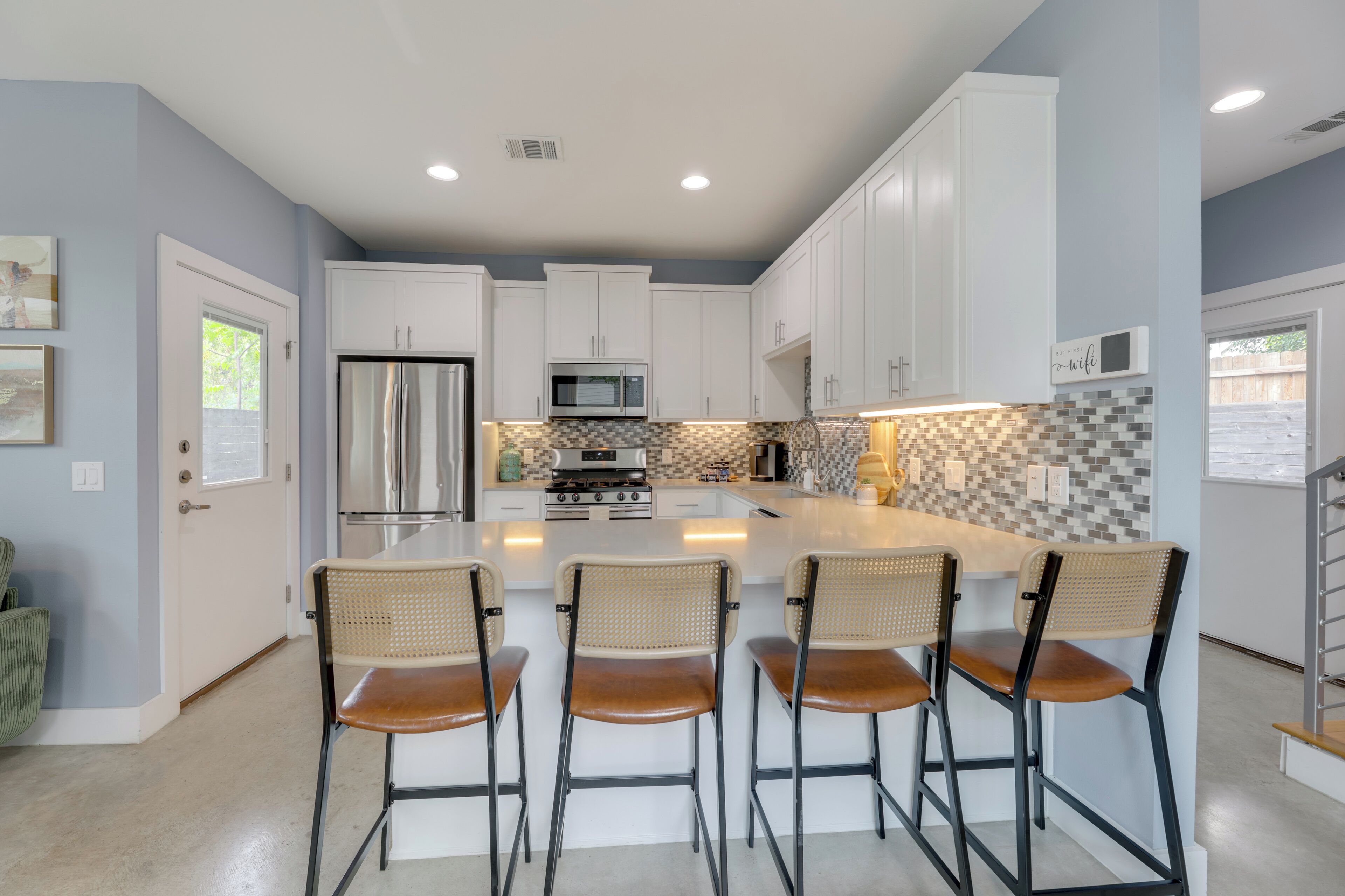 A modern kitchen with white cabinets, a stainless steel refrigerator, a microwave, and a tiled backsplash. There are four bar stools with woven seats.