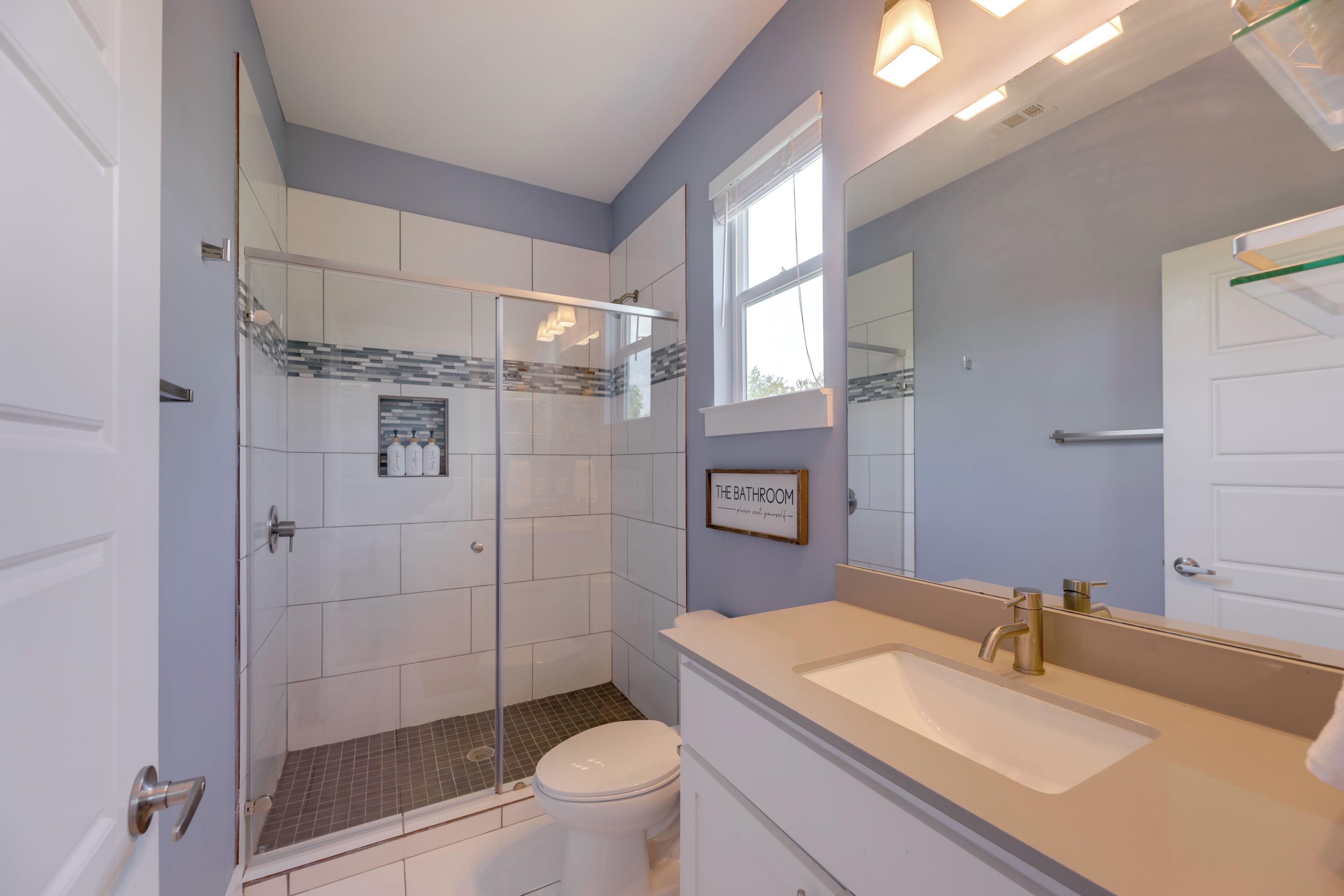 Een badkamer met een douchecabine van glas, een wit toilet, een wastafel met spiegel en een bordje met de tekst 'THE BATHROOM alstublieft jezelf instellen'.