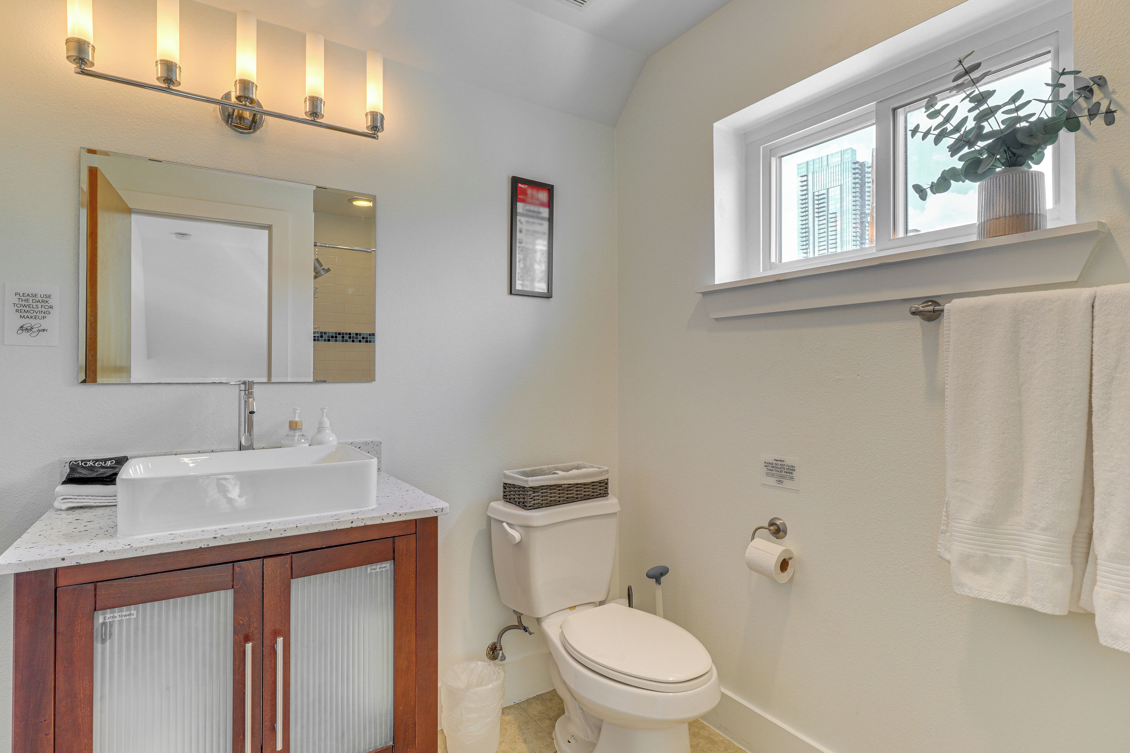 A bathroom with a white sink, mirror, and toilet. There is a window with a plant on the windowsill and a towel hanging on the wall.