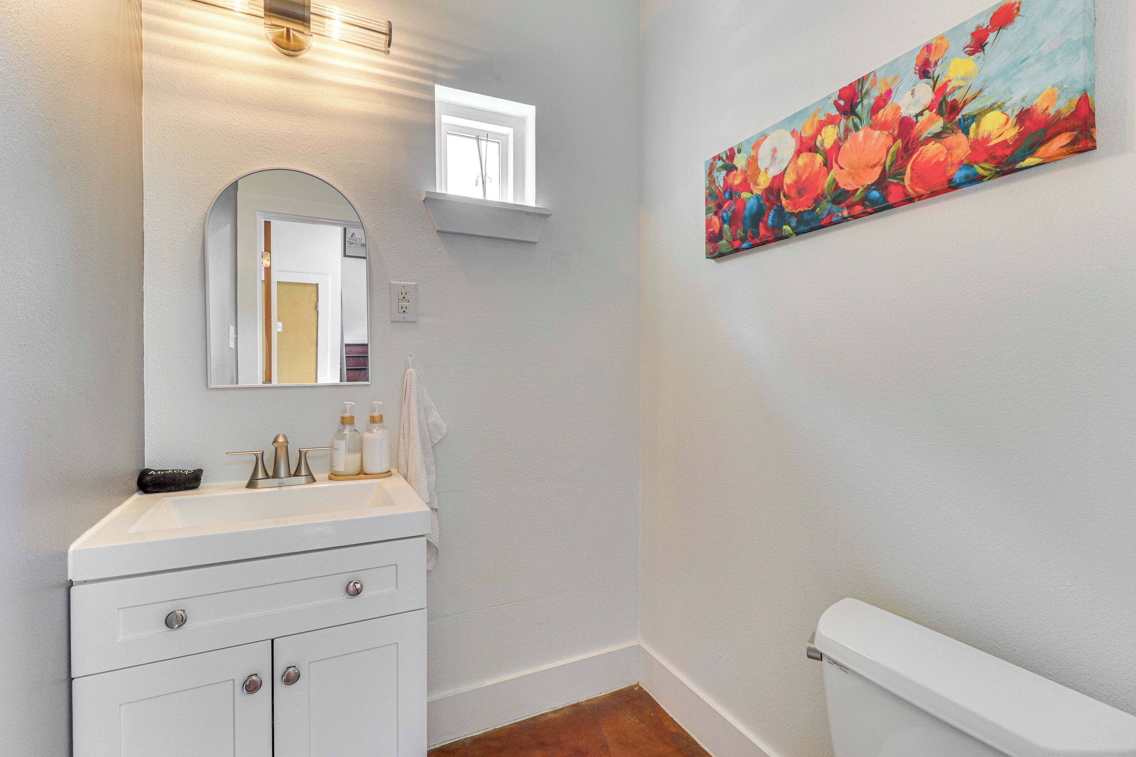 A bathroom with a white sink, a mirror, a painting of flowers, and a toilet.