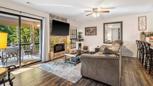 A spacious living room with a grey sofa, a coffee table, and a TV mounted on the wall. There is a balcony with outdoor furniture and a view of trees. - Cute, Comfy & Cozy - For 6 (Branson)