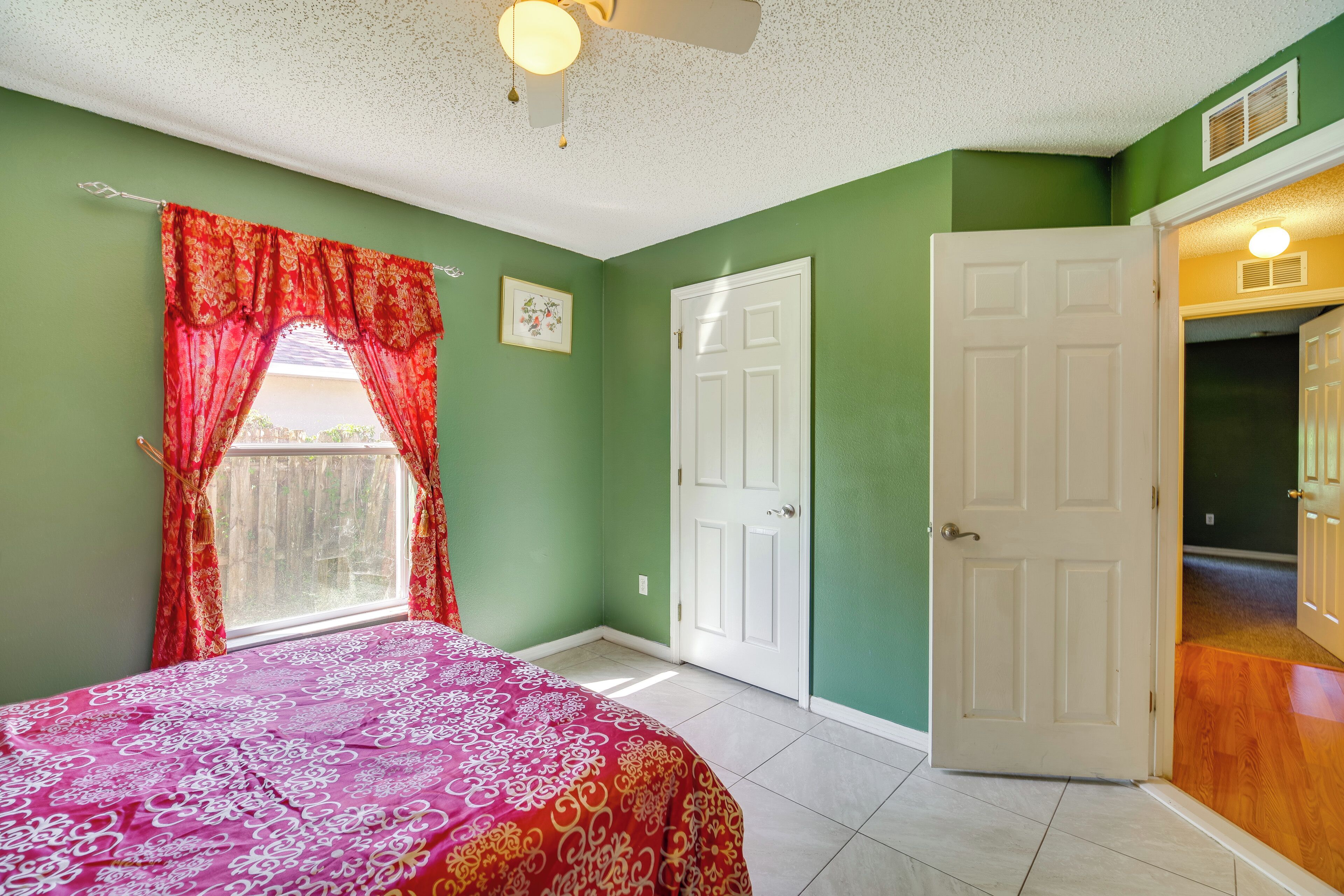 A bedroom with a bed, two doors, a window with red curtains, and a ceiling fan.