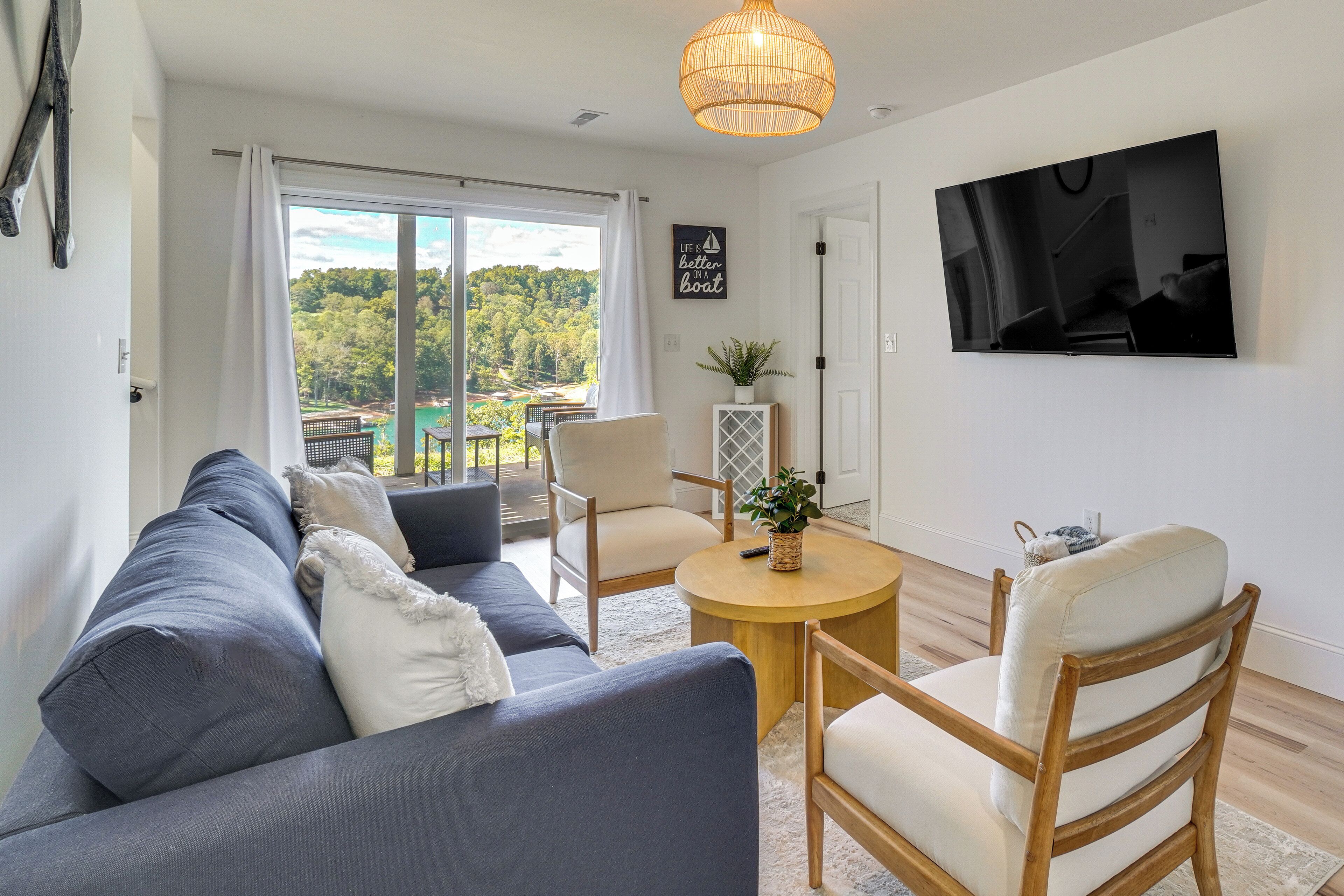A living room with a sofa, chairs, a round table, a TV, and a view of a green landscape through large windows.