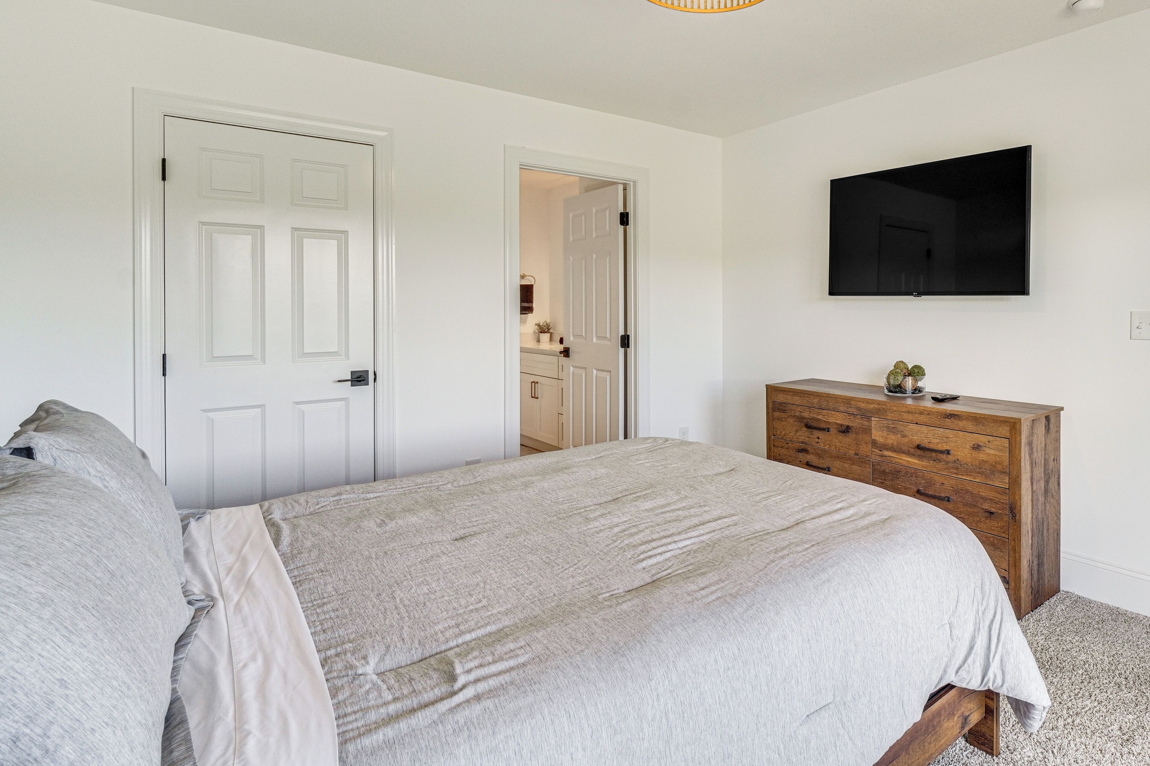 Une chambre à coucher avec un lit, une commode en bois, un téléviseur fixé au mur et une porte donnant sur une autre pièce.