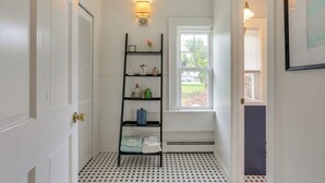A bathroom with a black and white tiled floor, a white door, a black shelf with toiletries, a window with a view of greenery, and a wall-mounted light fixture. - Walk to Hudson River Waterfront: Newburgh Home (Newburgh)
