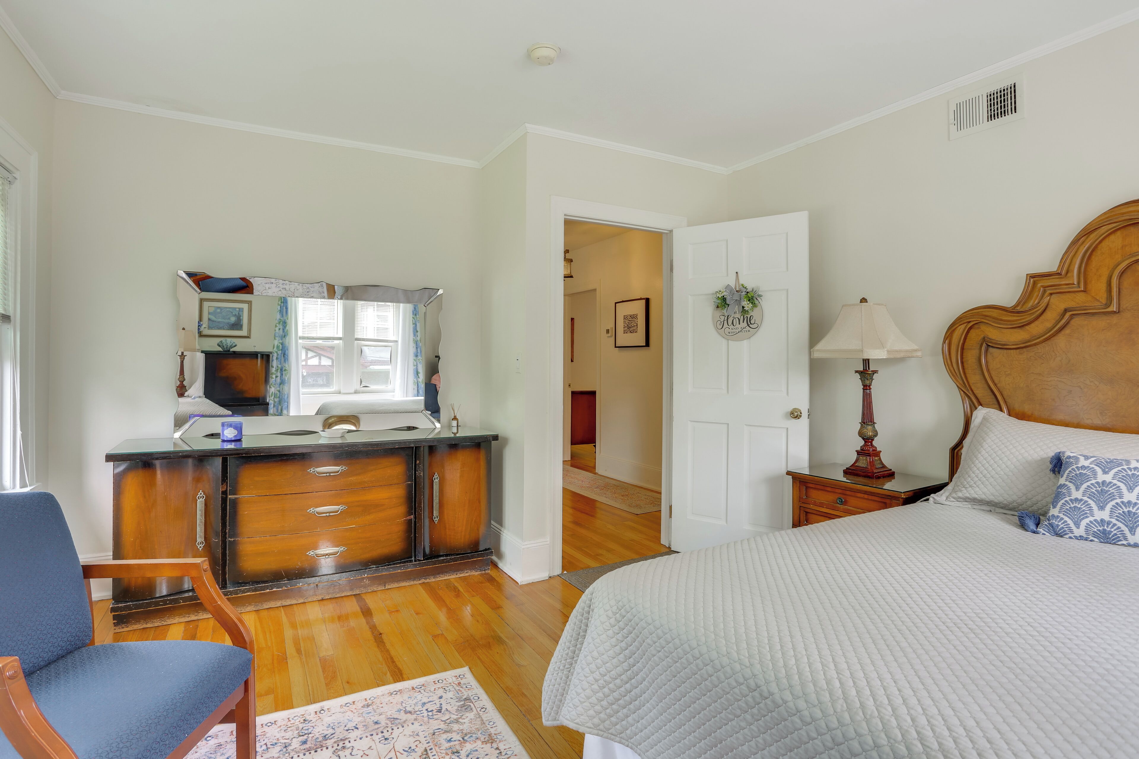Een slaapkamer met een bed, een stoel, een dressoir, een spiegel en een deur die naar een andere kamer leidt.
