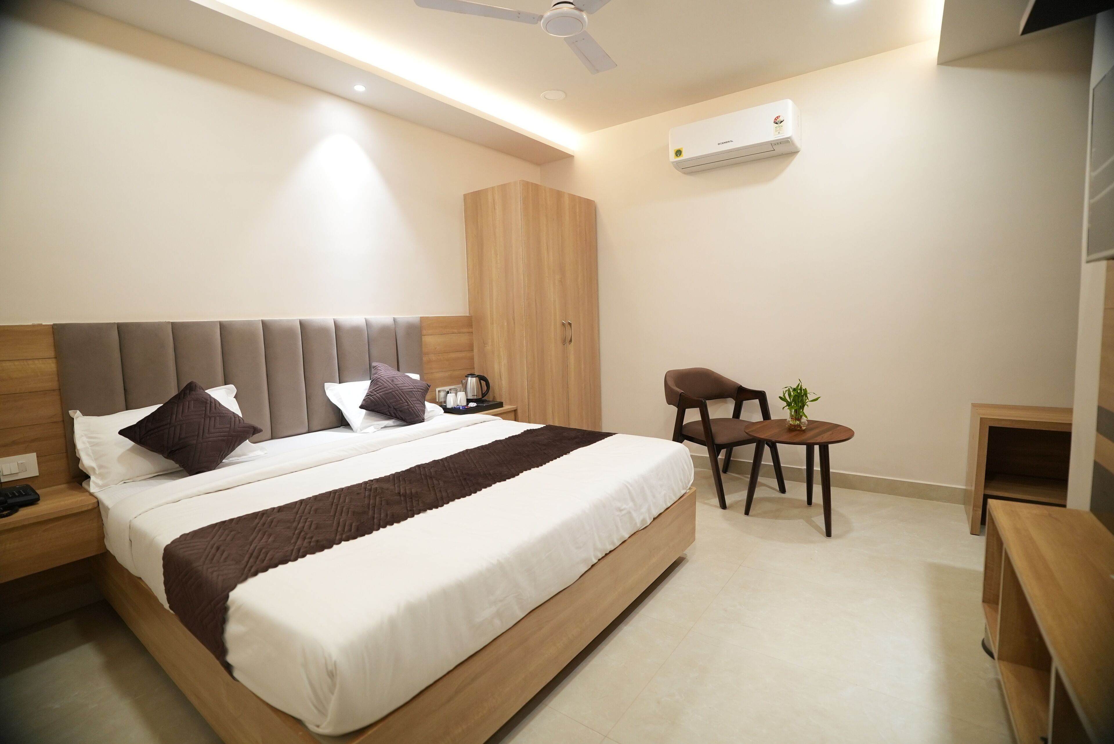 Ein modernes Hotelzimmer mit einem großen Bett, einem Holzschrank, einem kleinen Tisch mit Pflanze und einem Deckenventilator.