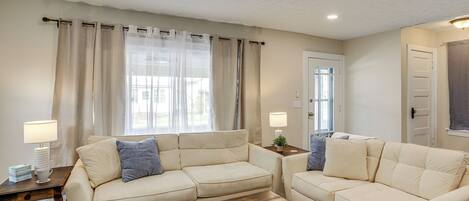 A living room with two cream-colored sofas, a wooden coffee table, and a window with sheer curtains.