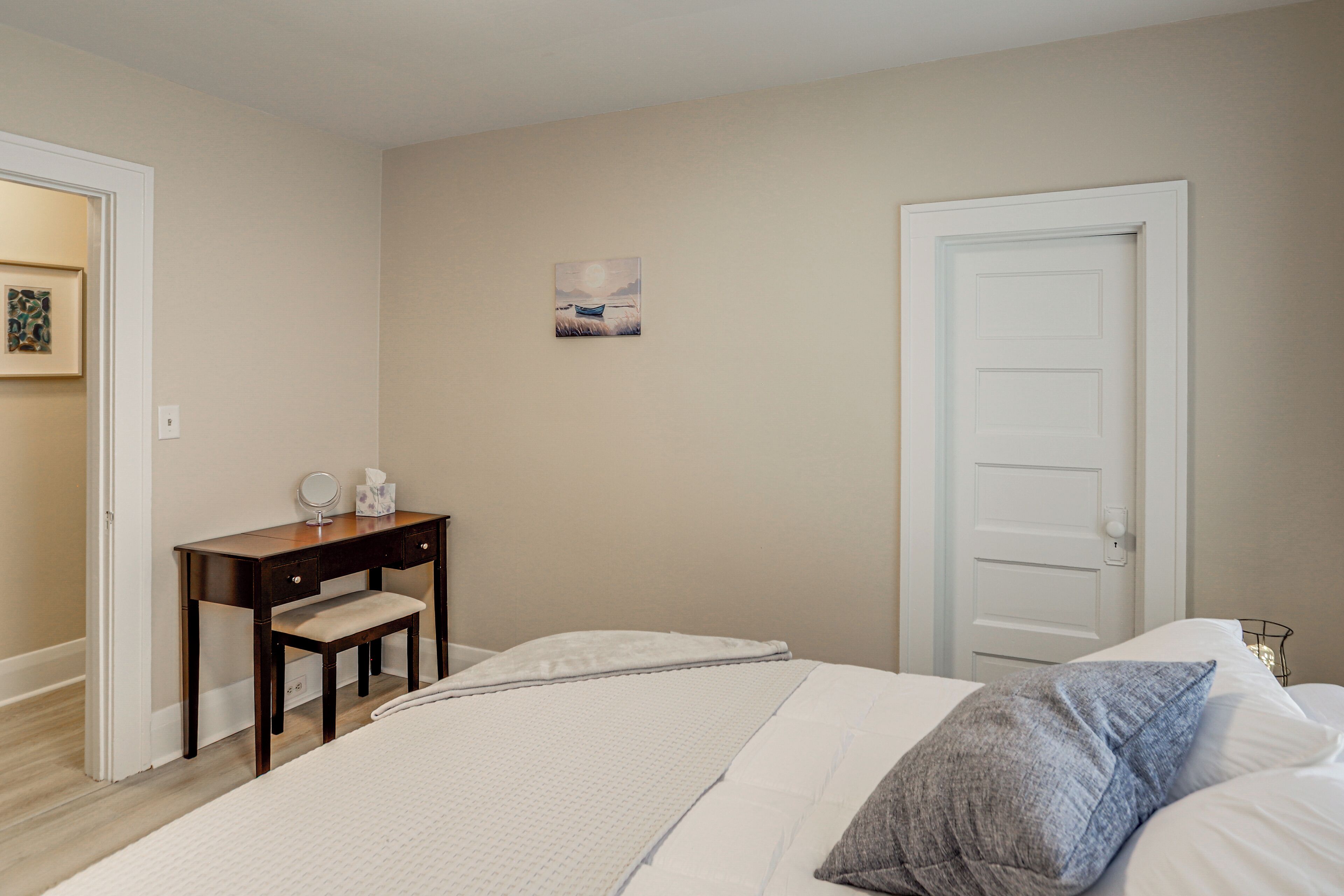 Ein Schlafzimmer mit Bett, Schreibtisch und Stuhl. Eine Tür und eine Wand mit einem Bild sind ebenfalls vorhanden.