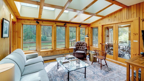 A spacious room with wooden walls, large windows, a leather armchair, a glass coffee table, and a wooden table. - Secluded Family Getaway w/ On-site Creek in Hawley (Charlemont)