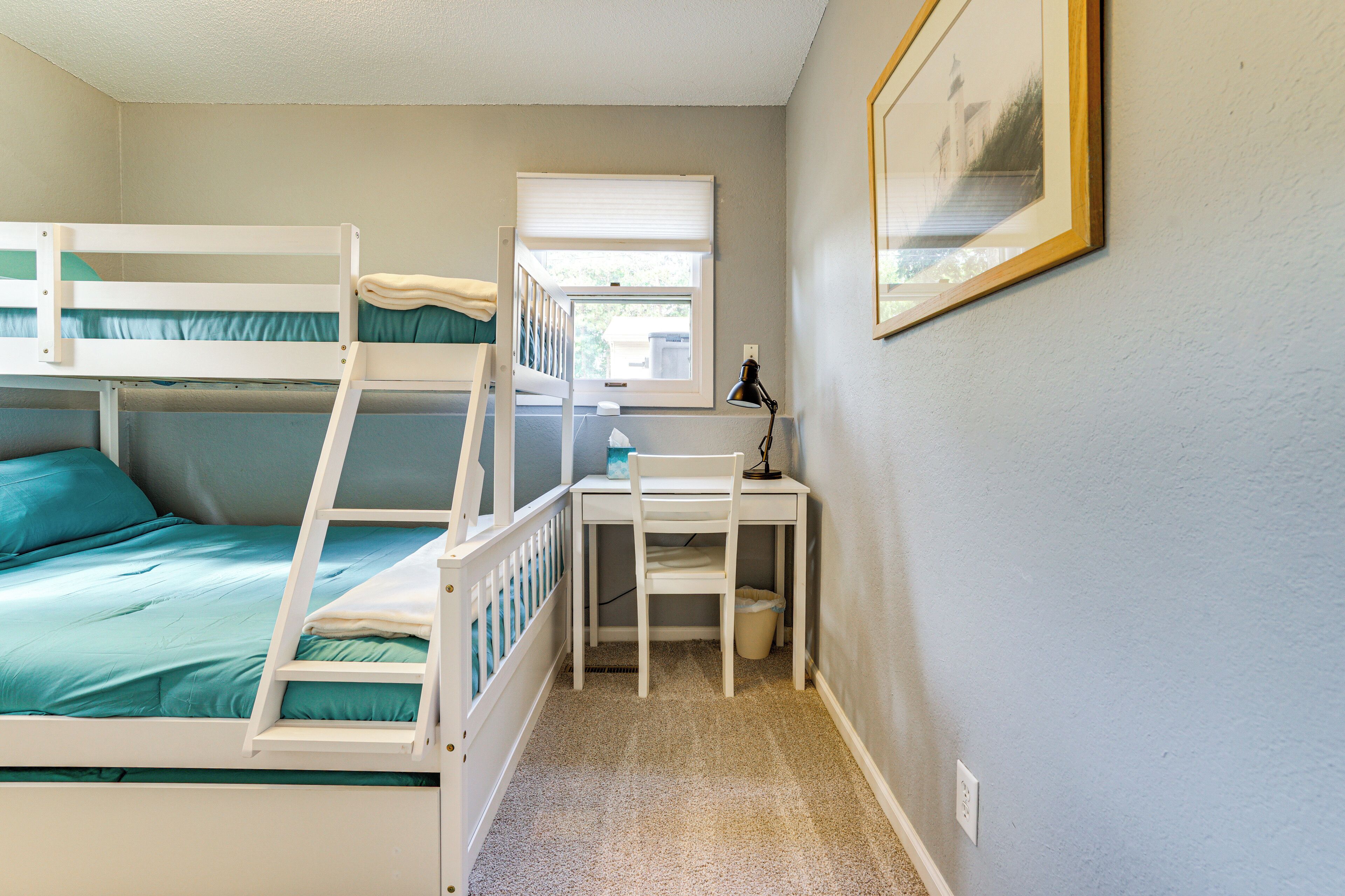 A room with bunk beds, a desk, a chair, a window, and a framed picture on the wall.