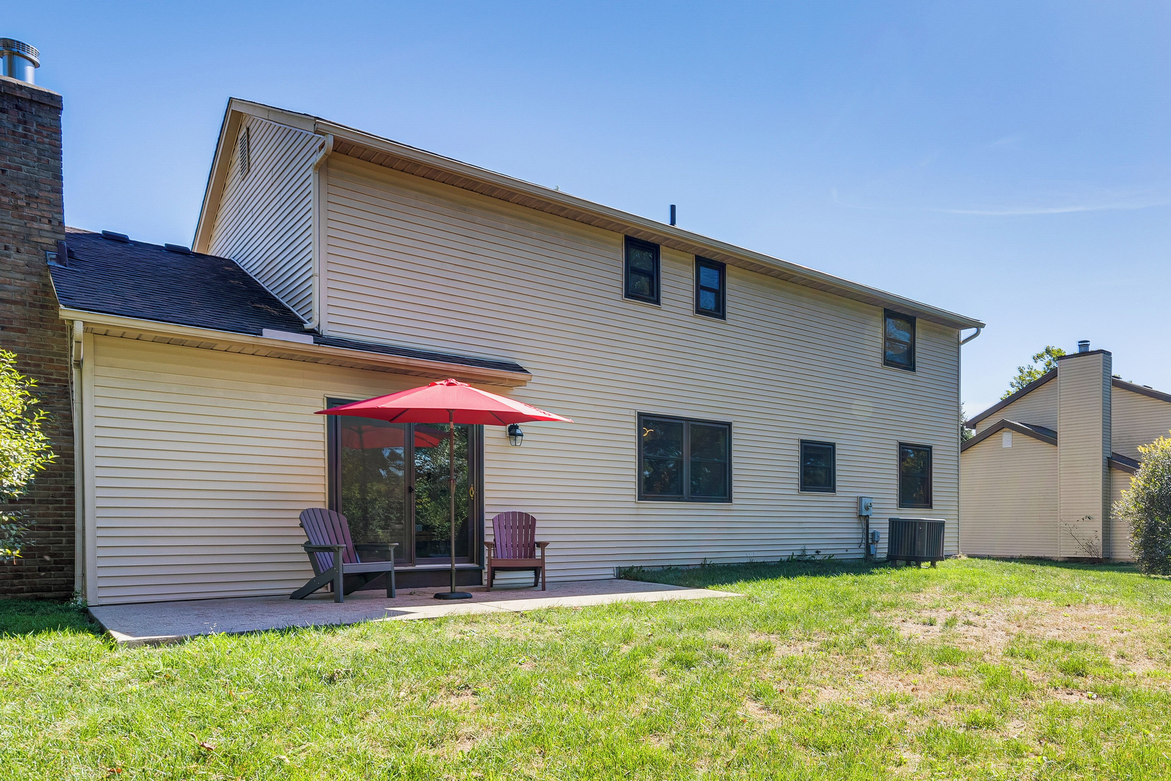 A two-story house with a covered patio area featuring two Adirondack chairs and a red umbrella.