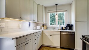 A modern kitchen with white cabinetry, a stainless steel dishwasher, and a window providing natural light. - Marinafront Cottage Sunrise, Fire Pit, Fireworks (Cambridge)