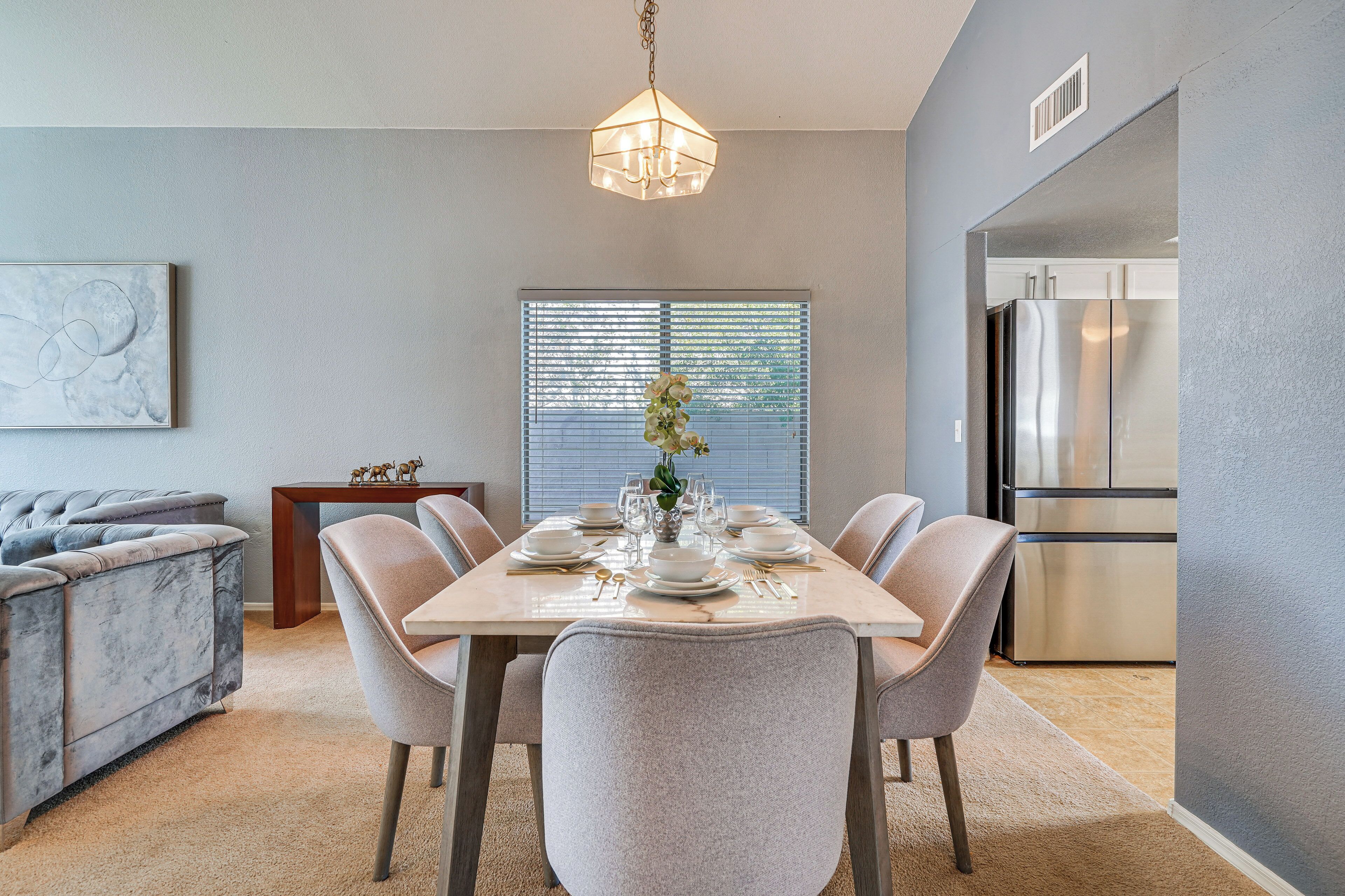 Uma sala de jantar com uma mesa posta para seis lugares, um sofá cinza, um candelabro e uma cozinha com uma geladeira de aço inoxidável.
